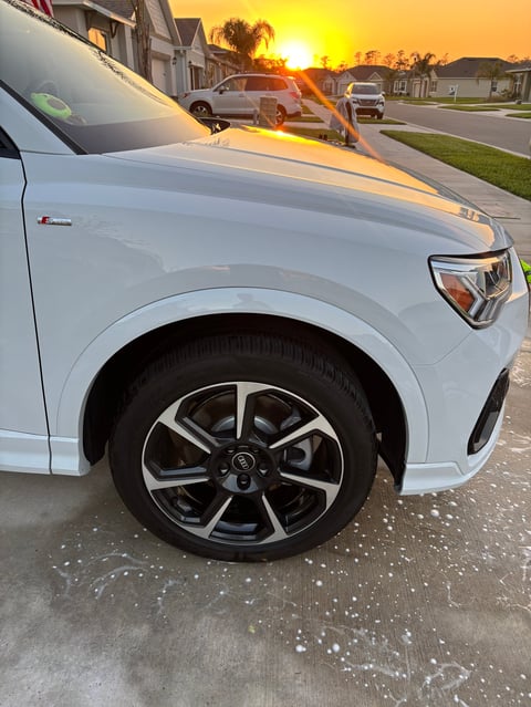 White SUV parked on residential driveway at sunset, showing front wheel and hood detail with palm trees and houses in background